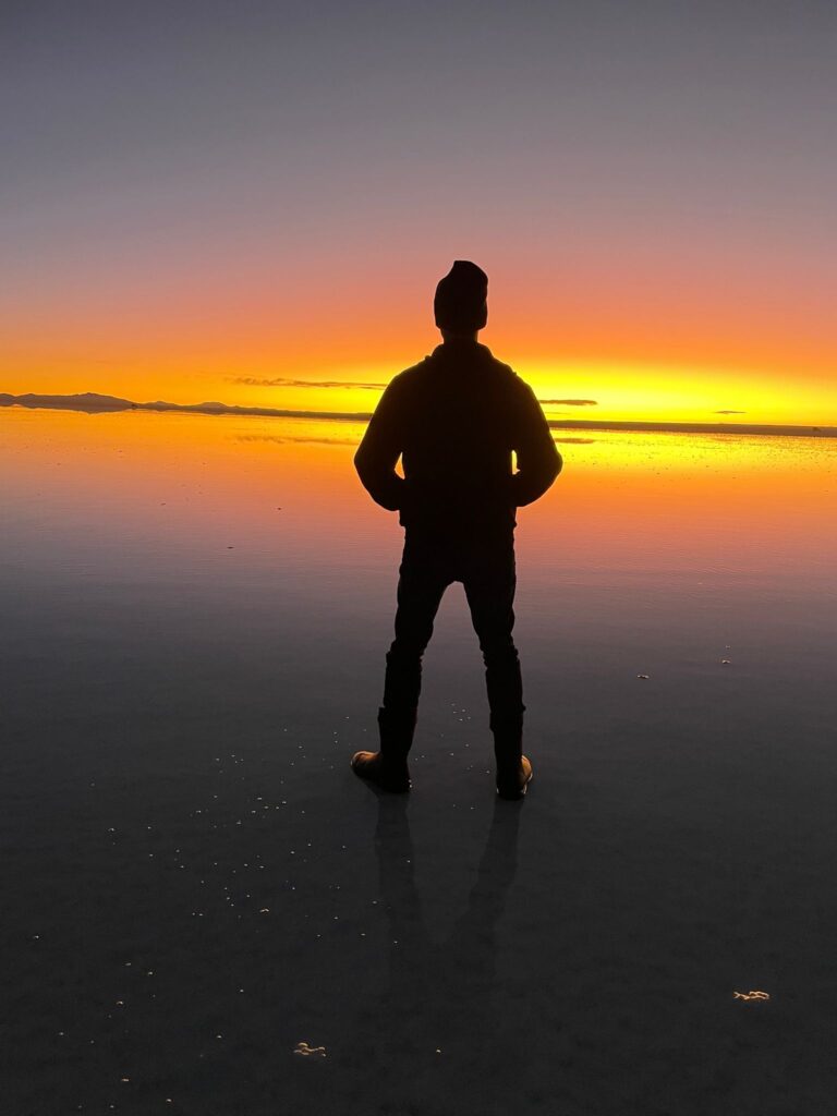 A picture of myself at Salar de Uyuni in Bolivia. I look like a silhouette standing on the dark salt, with the only light being a bright orange streak across the middle of the image, which comes from the sun setting