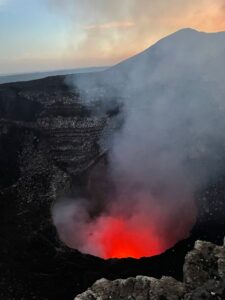 Lava inside Volcan Masaya in Nicaragua. You can see bright orange lava inside the circular crater, with thick grey smoke rising out of it. The orange sunset can just about be seen in the background, although it's tricky to fully make out with all the smoke in the way