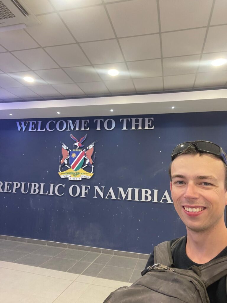 Alex standing by a sign that says "Welcome to the Republic of Namibia" to celebrate entering his 100th country.