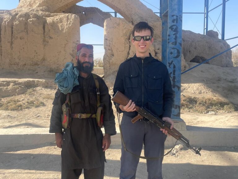 Alex standing alongside his Taliban bodyguard in Balkh, Afghanistan, whilst holding the guard's gun.