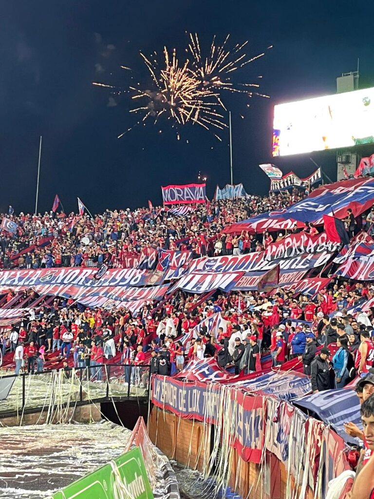 Football fans creating an atmosphere at an Independiente Medellin game in Colombia. They are holding banners supporting their team, whilst fireworks go off overhead,