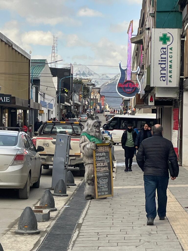 A man wearing a fluffy monster suit holds a sign promoting his chocolate and churro shop in Ushuaia, Argentina. He stands in the street where you can see several buildings including a pharmacy and Hard Rock Cafe. You can see snowy mountains visible in the distance.