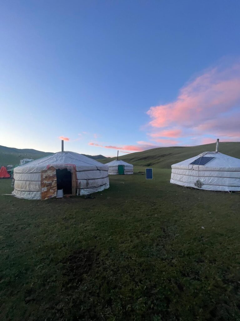 An image of 3 gers (yurts) in the Mongolian countryside, with red clouds in the background as the sun begins to set.