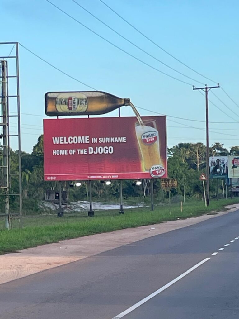 A sign near Paramaribo's airport that says "Welcome in Suriname, home of the DJOGO". There is an image of a djogo (a 1 litre bottle of Suriname's national beer, Parbo) being poured into a glass.