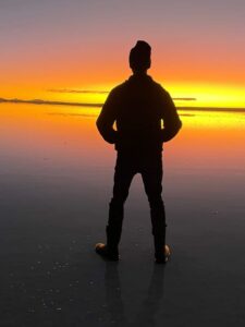 A picture of myself at Salar de Uyuni in Bolivia. I look like a silhouette standing on the dark salt, with the only light being a bright orange streak across the middle of the image, which comes from the sun setting