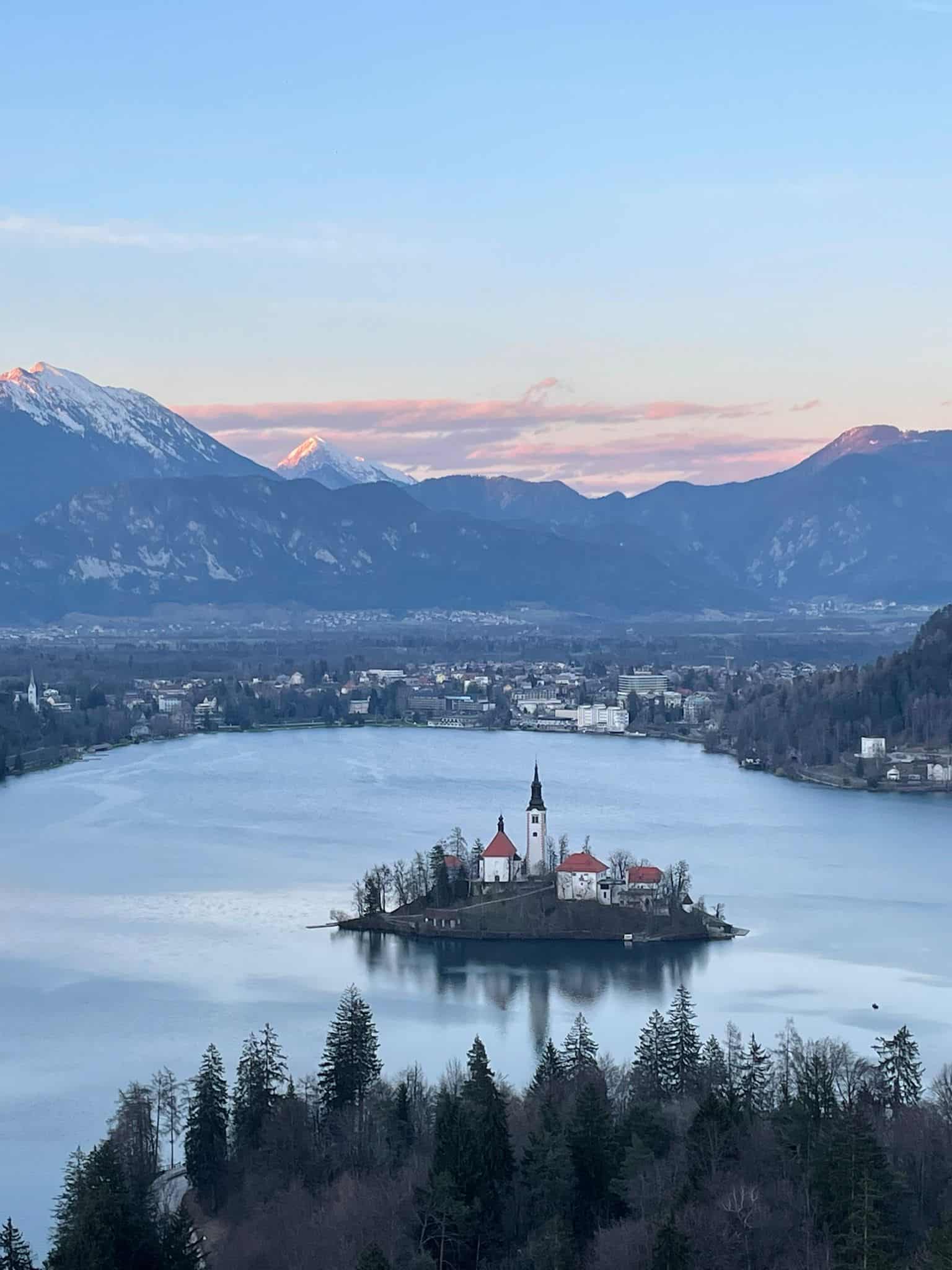 Bled Island standing in the middle of Lake Bled, Slovenia. The lake surrounds the island as the sun sets in the background behind snow-capped mountains.