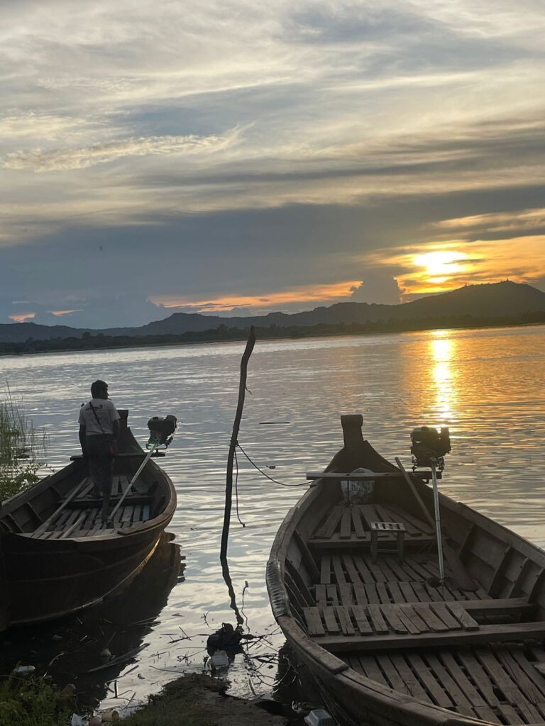 Two wooden boats on the Irrawaddy River in Bagan, Myanmar. A local stands on the left boat as the sun sets across the water