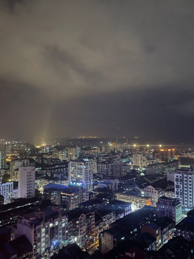 Yangon at night. You can see tall grey buildings spread across the city. There are several lights coming from all areas of the city.