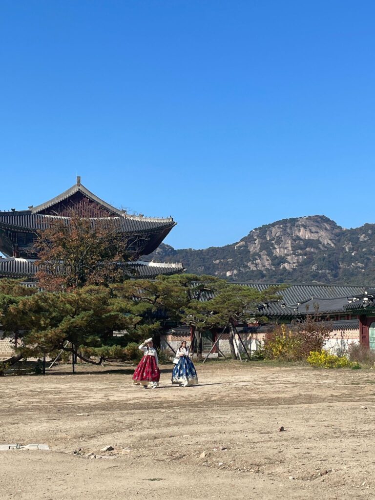 Two ladies wearing traditional South Korean hanboks at Gyeongbokgung Palace in Seoul. The lady on the left wears red whilst the lady on the right wears blue. They stand in front of a large traditional Korean building with trees in front and mountains in the background