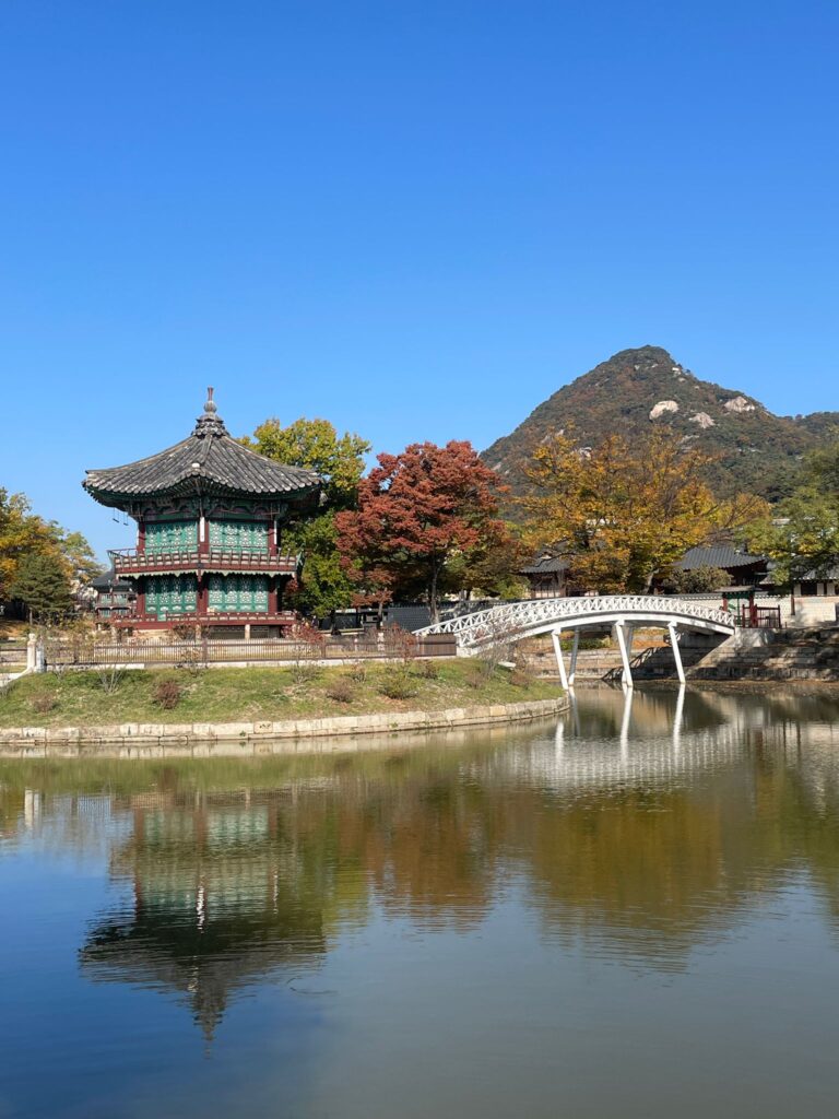 Hyangwonjeong Pavilion at Gyeongbokgung Palace in Seoul. The pavilion stands on a small island beside an artificial lake. A white bridge is to the right of it, with mountains in the background towering above the trees
