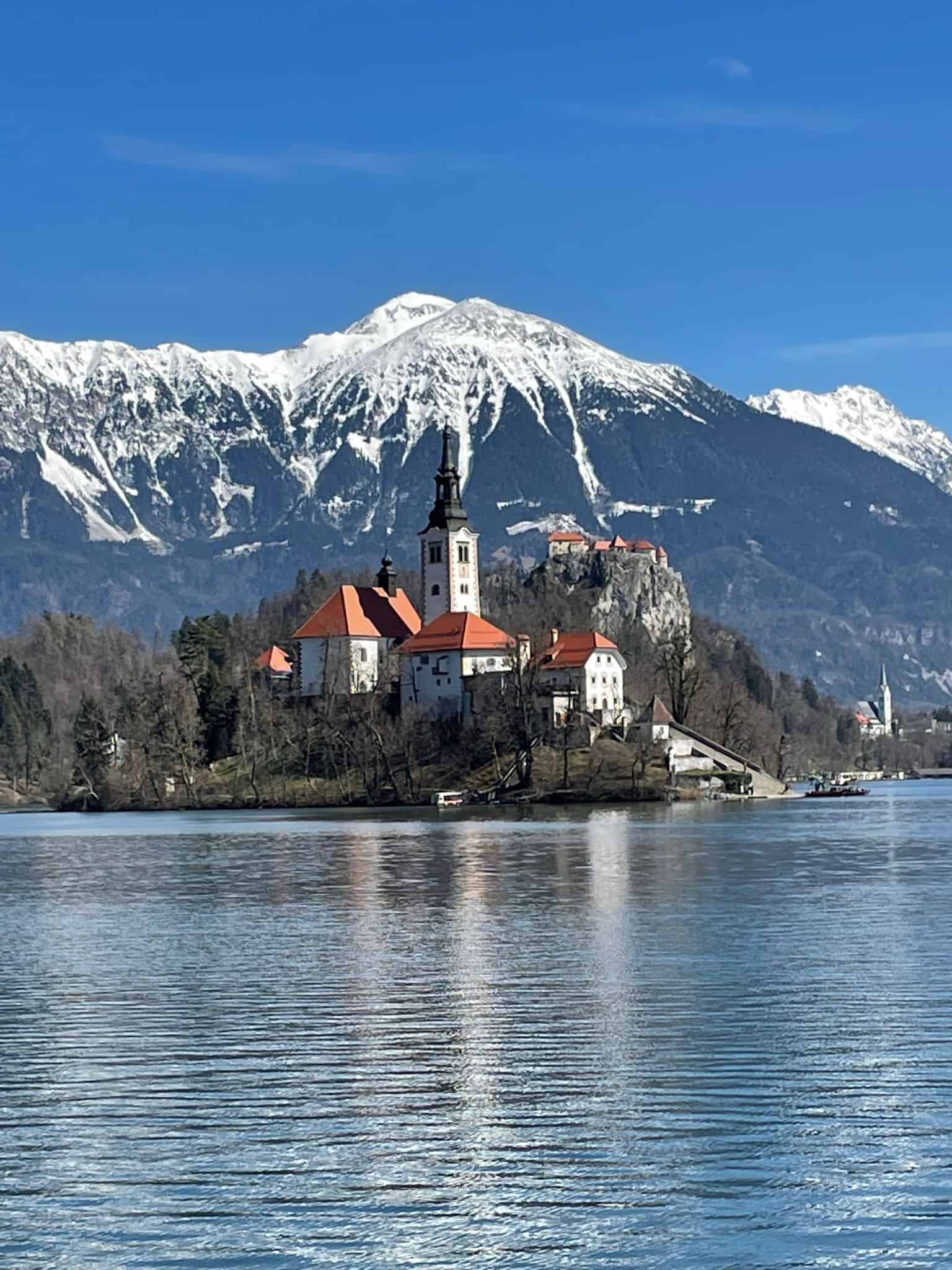 Bled Island in the middle of Slovenia's Lake Bled, where some buildings are located with snowy mountains surrounding the lake in the background.