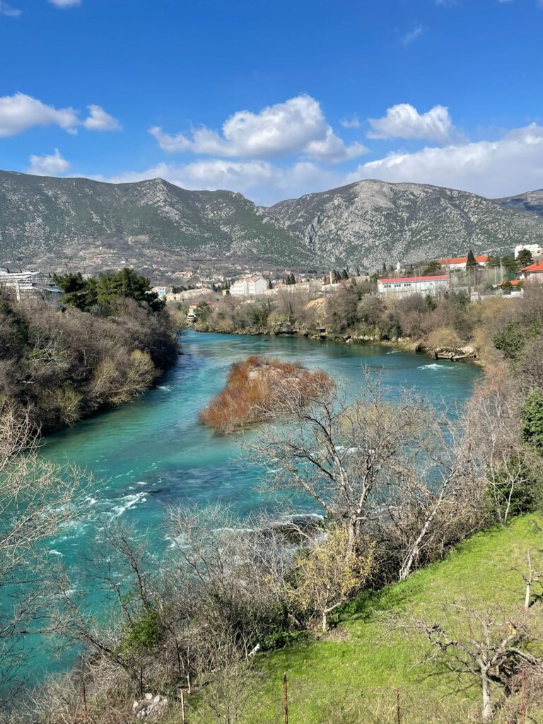 Views of the Neretva river featuring mountains in the background and the turquoise river separated by a small island. Picture taken from Carinski Most, a bridge in Mostar, Bosnia Herzegovina