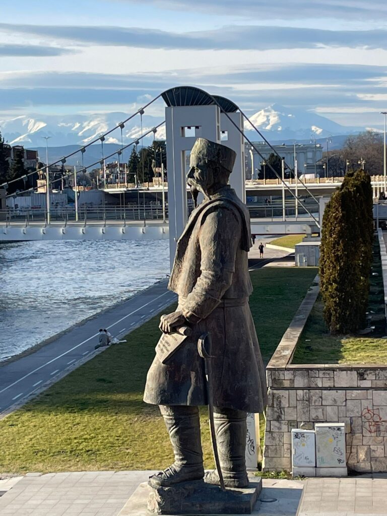 A statue in Skopje, North Macedonia, next to the Vardar River with a bridge and snow-capped mountains in the background