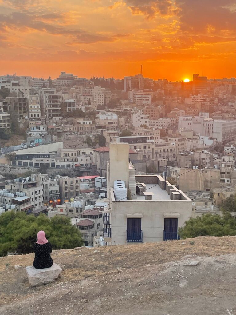 A local in a hijab with her back to the camera observing a bright sunset over Amman. The sun sets in the background over the city's buildings. Picture taken from Amman Citadel