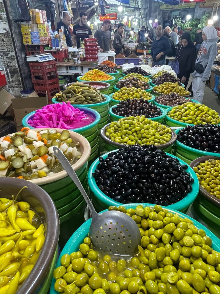 A series of colourful fruits and olives in Amman's Souq Al-Sukar, with several locals in the background looking on