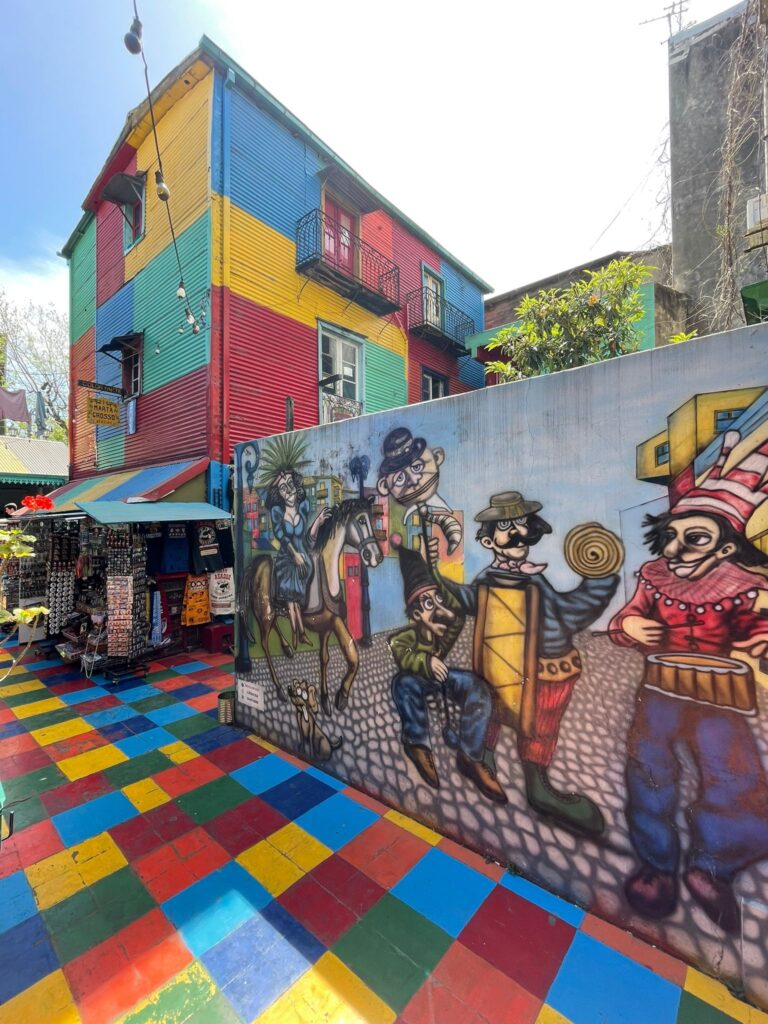 A colourful building in the famous street "El Caminito" in Buenos Aires. It towers over a wall displaying street art of some musicians, whilst a tourist souvenir stall stands in the background