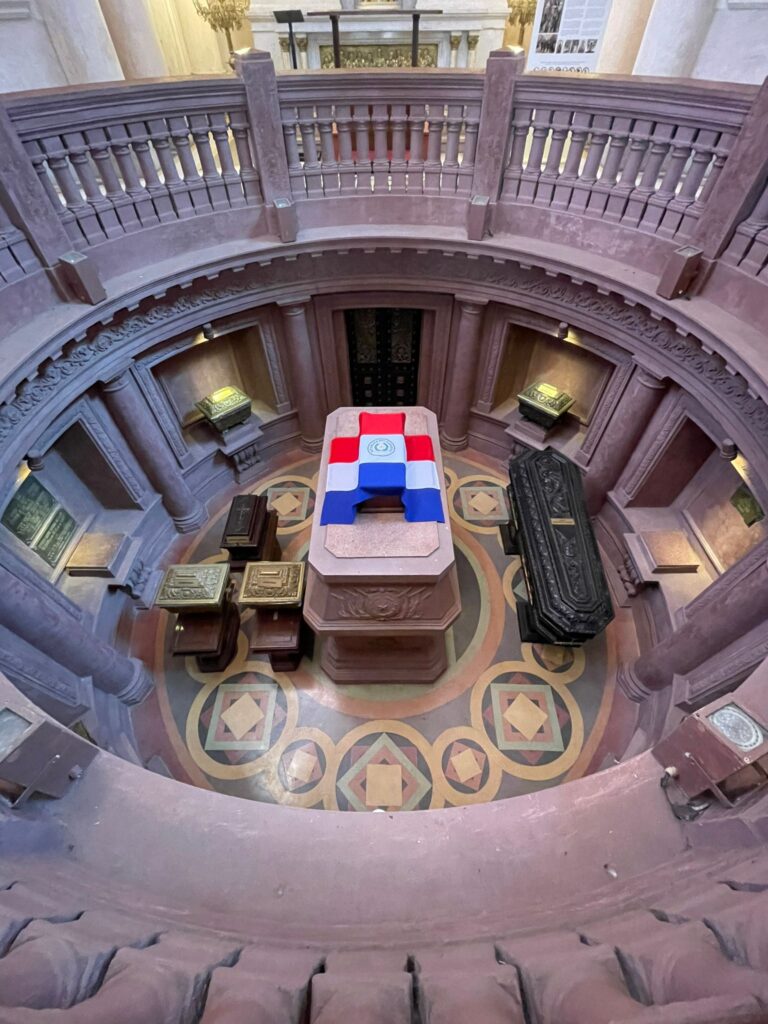 A series of coffins inside a pit in Asuncion's Pantheon of Heroes. In the middle is a large one covered in the Paraguayan flag. It is surrounded by many smaller coffins and urns