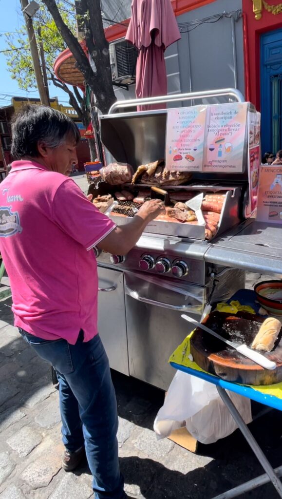 A man in a pink shirt cooking various meats on la parrilla at Resto Bar Las Gemelas in La Boca, Buenos Aires