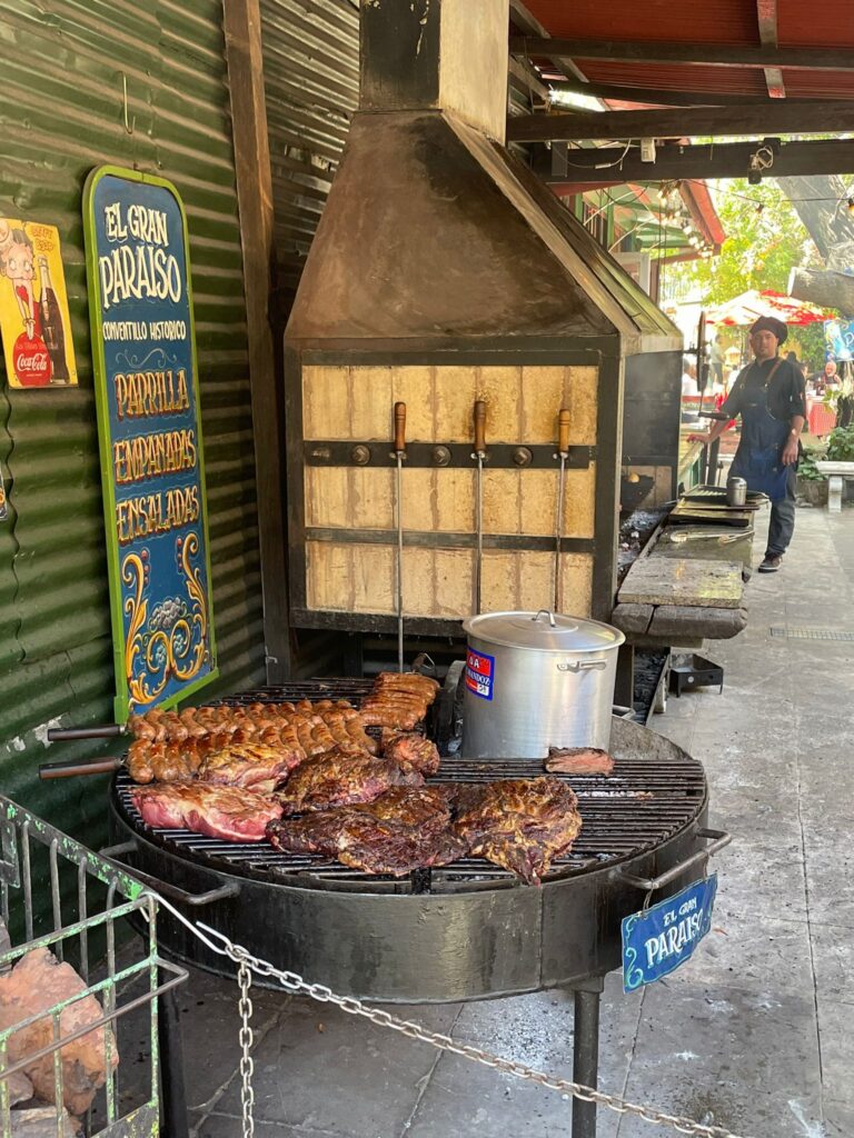 An Argentine Parrilla full of meats in La Boca, Buenos Aires. An asador stands in the background. There is a sign to the left of the grill which reads "parrilla, empanadas, ensaladas". If you know how to order steak in Argentina then this guy is guaranteed to give you a good cut!