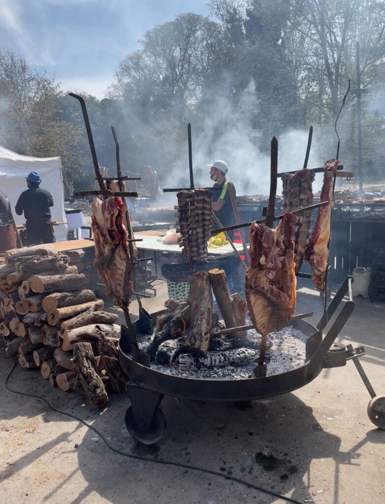 An asador grilling steak on the parrilla at a Chile-themed market in Mendoza. Various meats are being grilled on spits, with logs next to the parrilla to ensure it continues to cook the meat