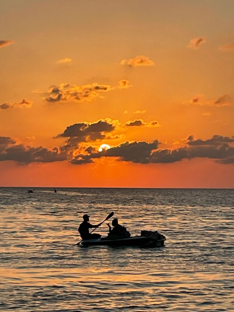 Two men in a small rowing boat just off the coast of Caye Caulker, Belize as the sunsets behind the clouds in the distance creating a bright orange sky