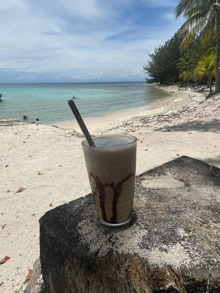 An image of a Monkey Lala cocktail with the yellow beach and blue waters of Utila in the background, alongside green trees