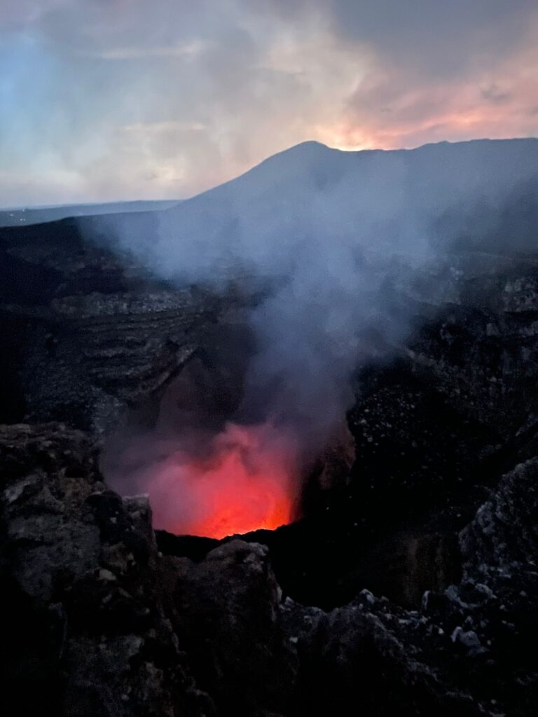 The Santiago crater of Volcan Masaya in Nicaragua. You can see orange lava with dark smoke rising out of the black volcano and obscuring the sunset views in the distance