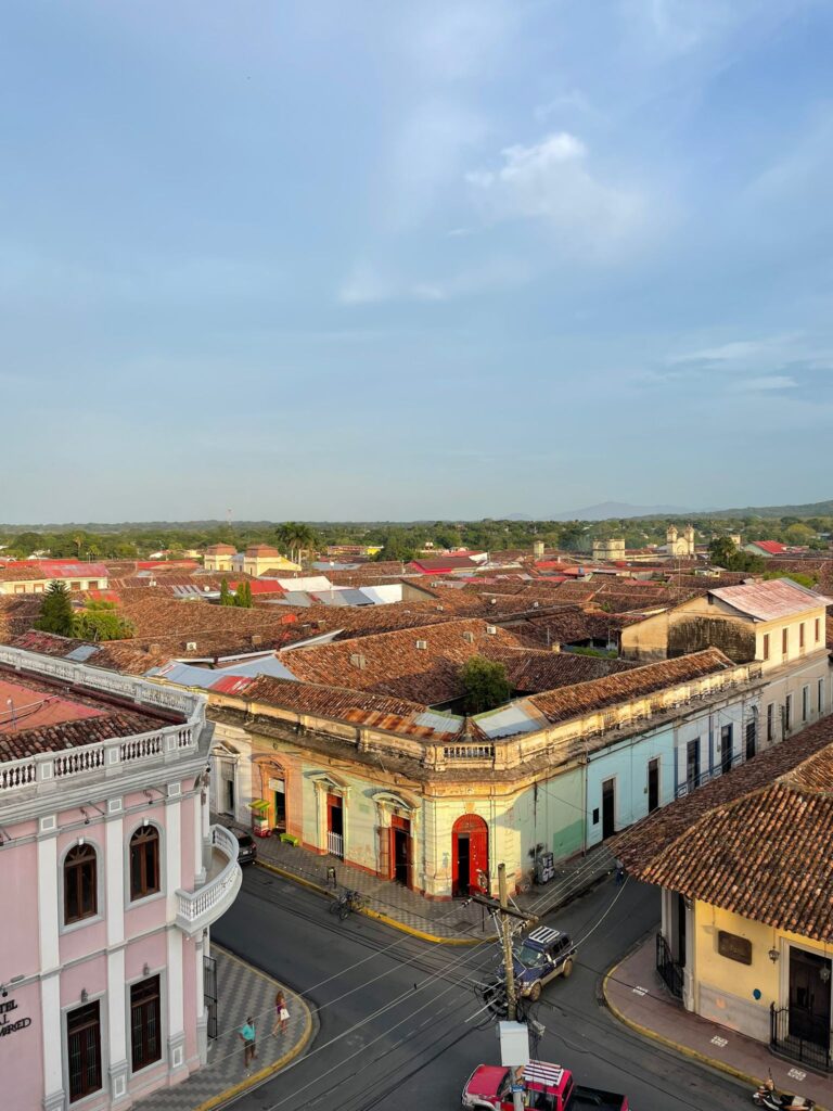 Several colorful colonial-style buildings in Granada, Nicaragua. From left to right you can see pink, white, pink, yellow, blue, white and yellow buildings. There is also a crossroads separating the first from the rest with electrical lines hanging overhead, and the faint image of a volcano in the background. Green trees separate the buildings from the volcano, whilst the skies are blue with few clouds to be seen 