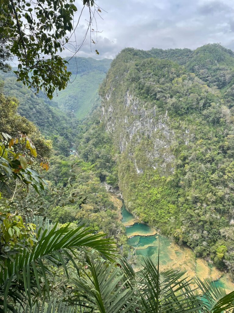 A series of turquoise pools in the shape of a naturally-formed "staircase" at Semuc Champey in Guatemala. The pools are surround by green trees from the surrounding rainforest with clouds descending in the background 