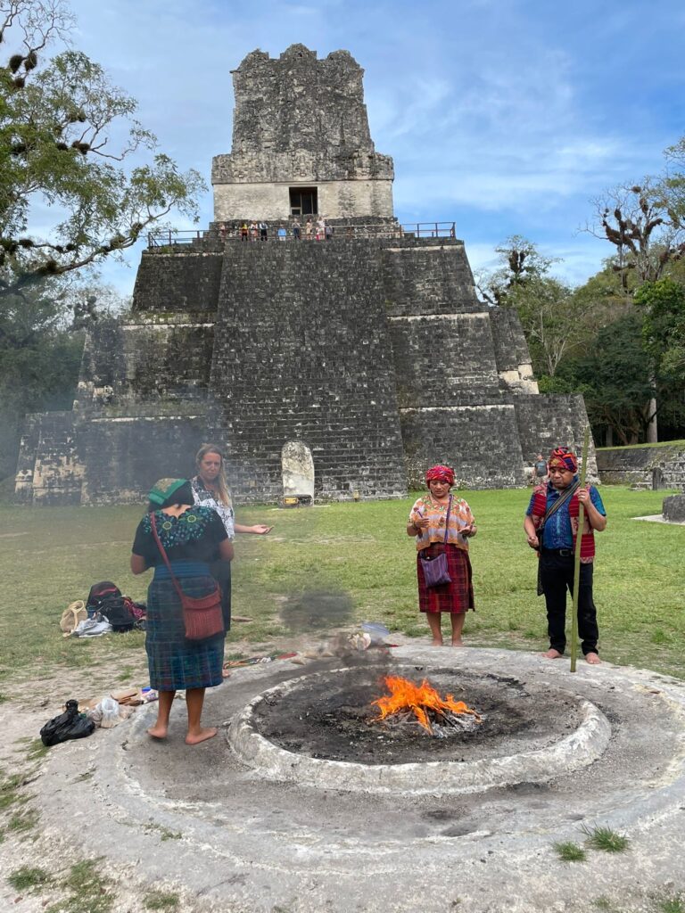 Three locals and a tourist participate in a Mayan ceremony around a bonfire whilst wearing traditional clothing. They are stood in front of the famous Tikal ruins