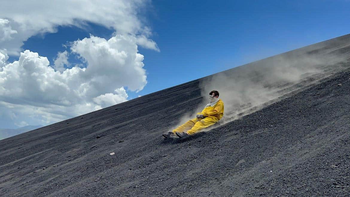 A photo of myself bombing down Cerro Negro Volcano in Nicaragua on a volcano board. I'm wearing a protective yellow suit as a large cloud of gravel flies up in the air behind me.