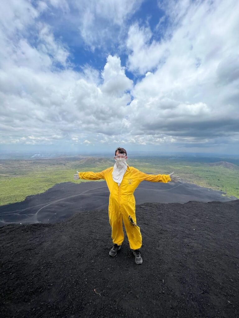 A picture of myself on top of Cerro Negro Volcano in full protective gear: a yellow jumpsuit, gloves, goggles and a white bandana. I also wear my own hiking boots whilst standing on the black ash at the top of the volcano. A must for any Nicaragua itinerary