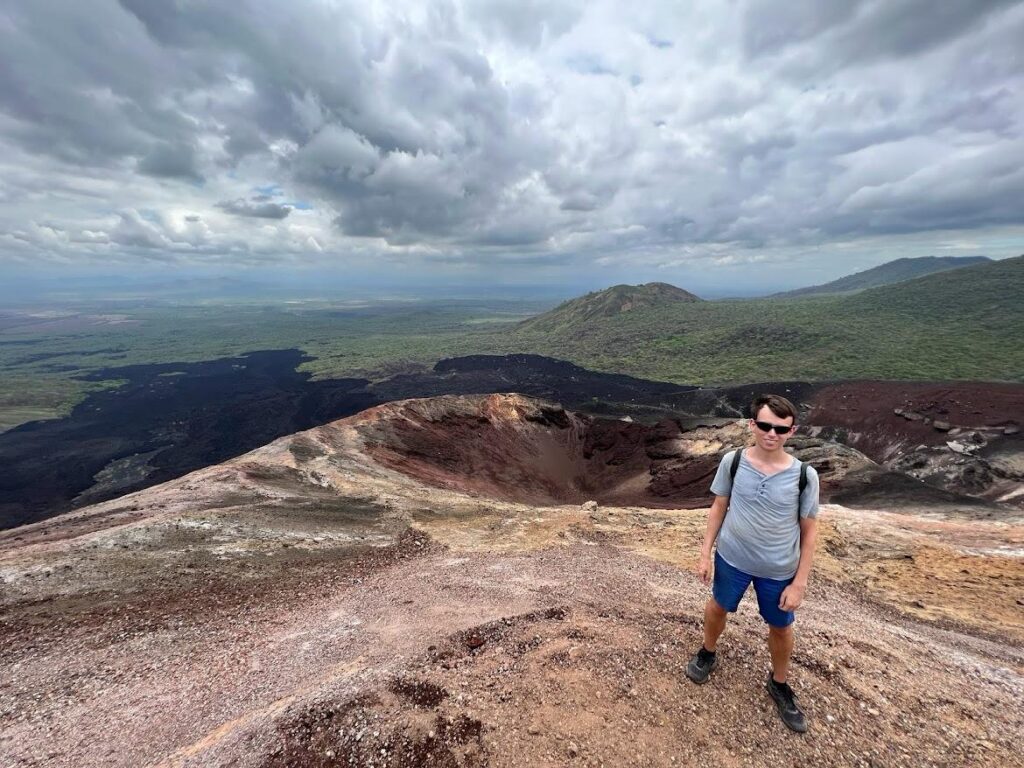 A picture of myself in blue shorts and a blue shirt standing at the summit of Cerro Negro Volcano in Nicaragua, with the crater behind me in the middle of the image