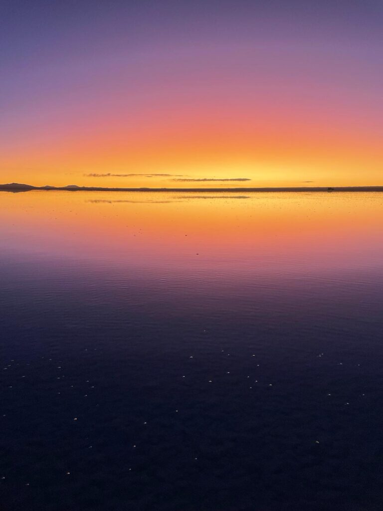 The best time to visit Salar de Uyuni in Bolivia is during the wet season as you increase your chances of being able to see the mirror effect as shown in this image. The picture shows bright orange skies at sunset with purple reflected at the top and bottom of the photos, and a symmetrical image of the dark clouds and mountains forming a thin line across the middle