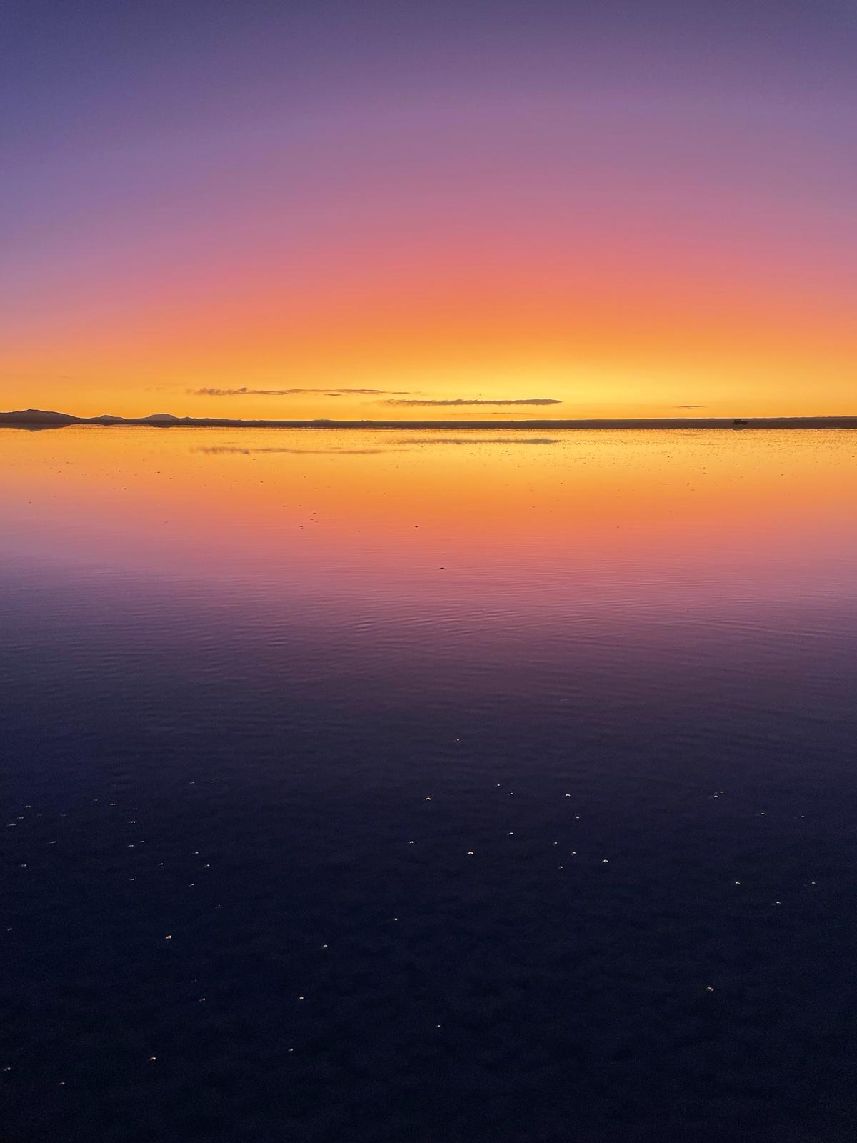 The best time to visit Salar de Uyunu in Bolivia is during the wet season as you increase your chances of being able to see the mirror effect as shown in this image. The picture shows bright orange skies at sunset with purple reflected at the top and bottom of the photos, and a symmetrical image of the dark clouds and mountains forming a thin line across the middle.