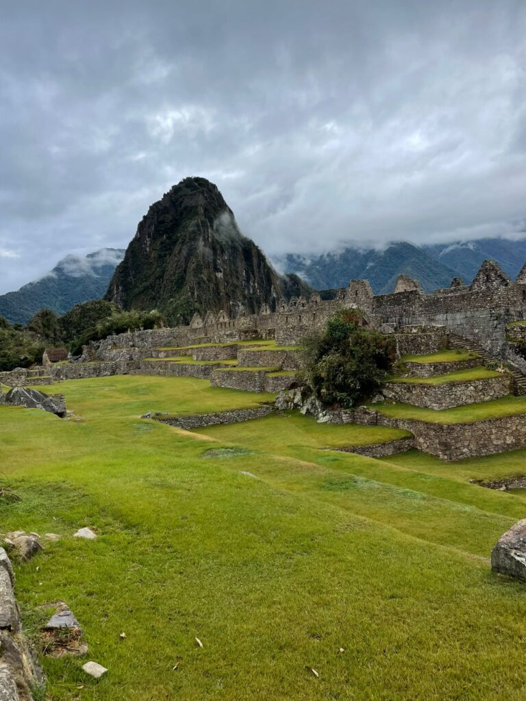 A mountain standing above the ruins of Machu Picchu at the famous Inca site, with clouds in the background floating across the grey skies
