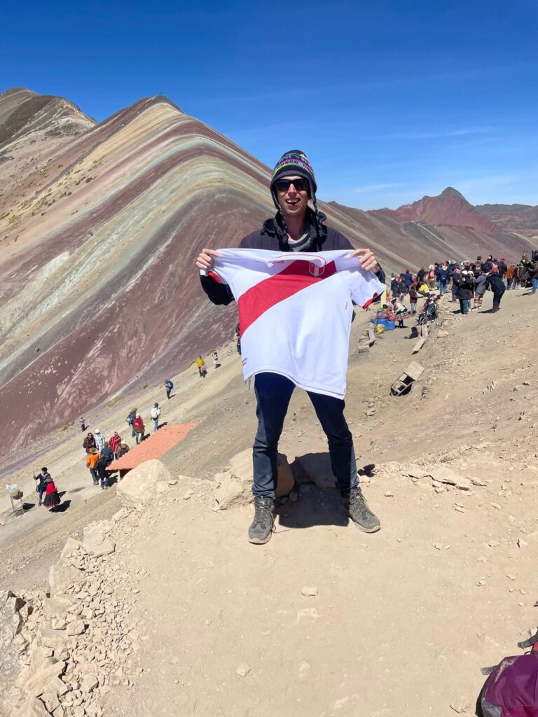 A picture of me holding a Peru football shirt at the top of Rainbow Mountain. The mountain is a mixture of a reddish-brown colour along with more yellowish colours and lighter shades of brown