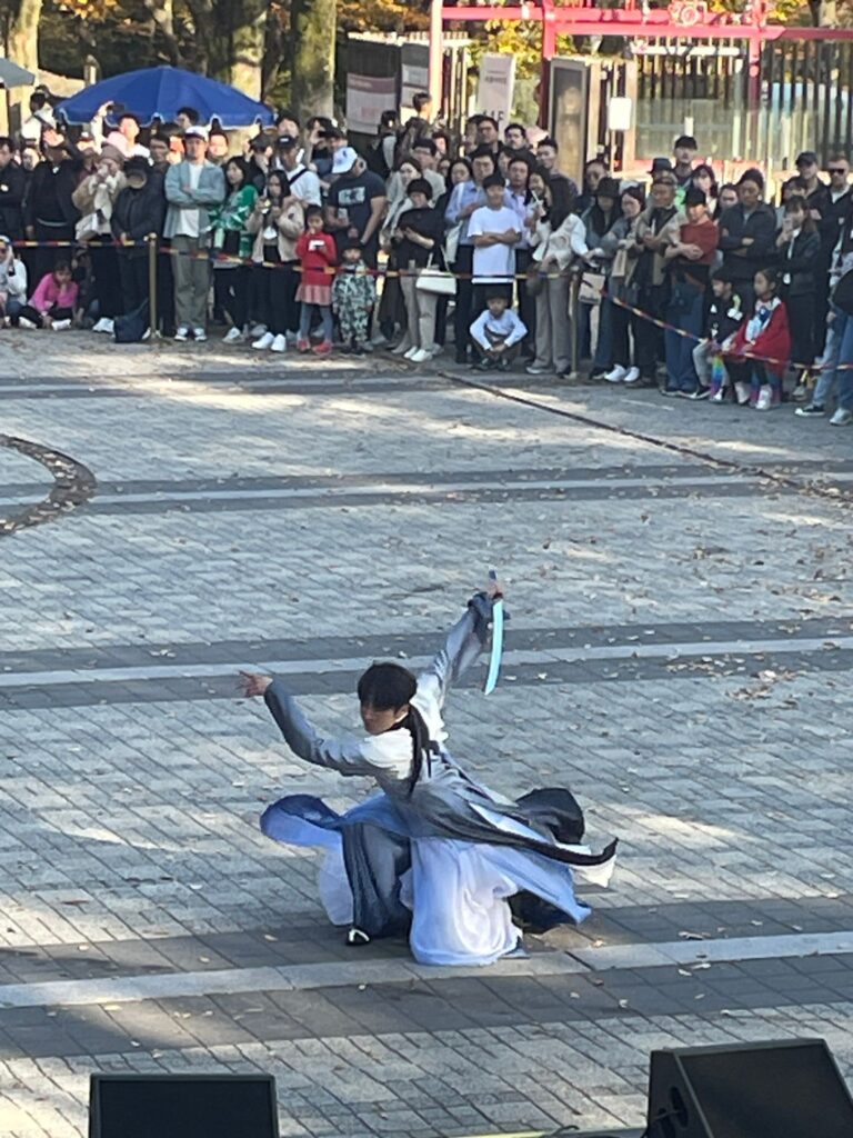 A man in traditional Korean clothing with a large sword, putting on a martial arts performance for hundreds of people looking on at the base of N Seoul Tower in the capital