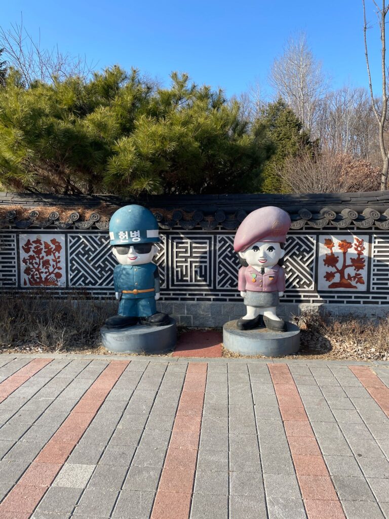 Statues of two guards, just like those at the JSA. On the left is a soldier dressed in blue with sunglasses on. On the right is a lady in a pink formal uniform