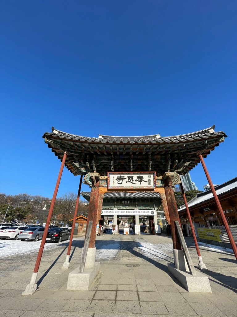 The entrance to Bongeunsa Temple in Seoul. It shows an archway in the traditional Korean style, being held up by wooden poles, with a white sign in the centre on which letters are written in the Korean language
