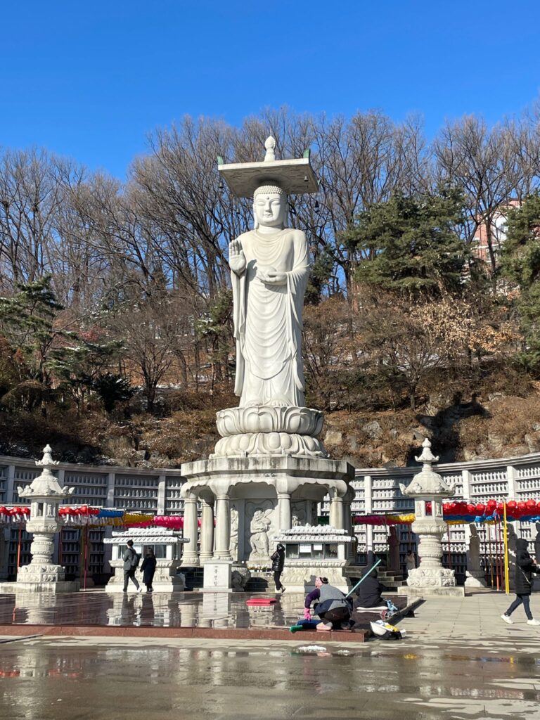 A tall white statue of a Buddha surrounded by thousands of tiny Buddhist statues lined up along the walls surrounding the big statue. This is at Bongeunsa Temple: One of the best places to visit even if you only have one day in Seoul