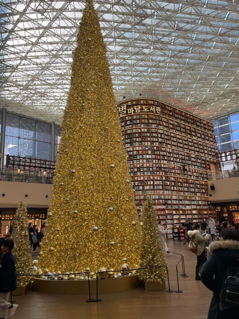 A golden Christmas tree reaching up to the ceiling in Seoul's Starfield Library. Books are stacked high in the background on neatly-arranged shelves with a sign written in Hangul at the top