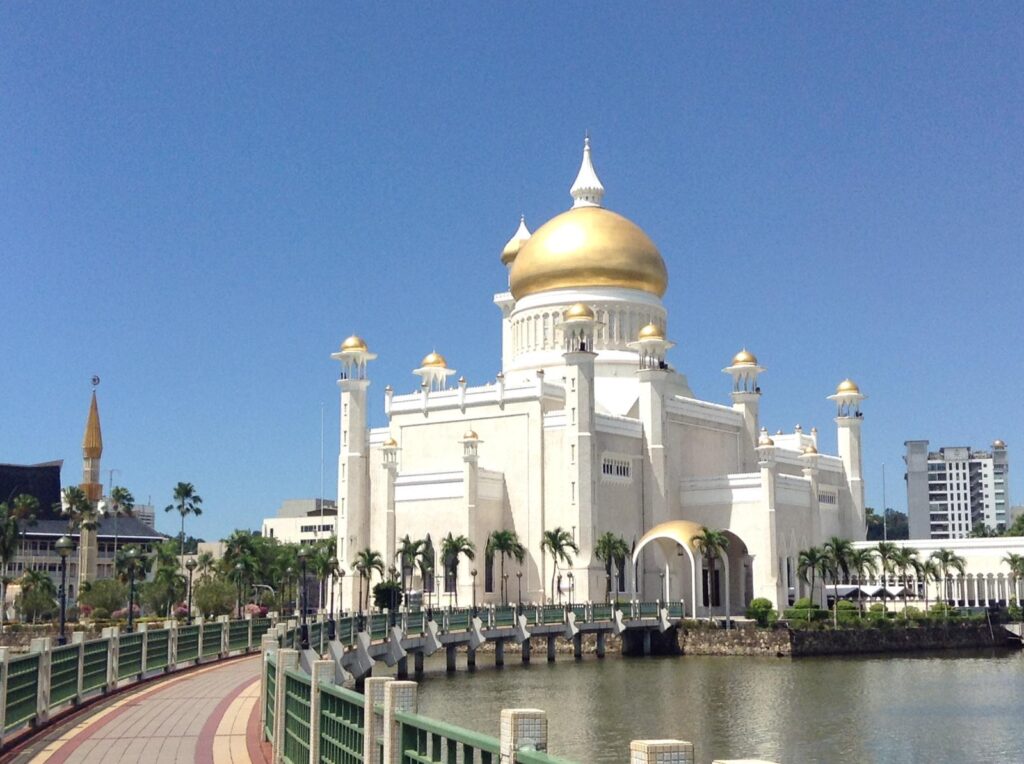 The gold-domed Omar Ali Saifuddien Mosque in Brunei. There is a curved walkway on the left lined with palm trees that connects the mainland with the mosque over a small lake
