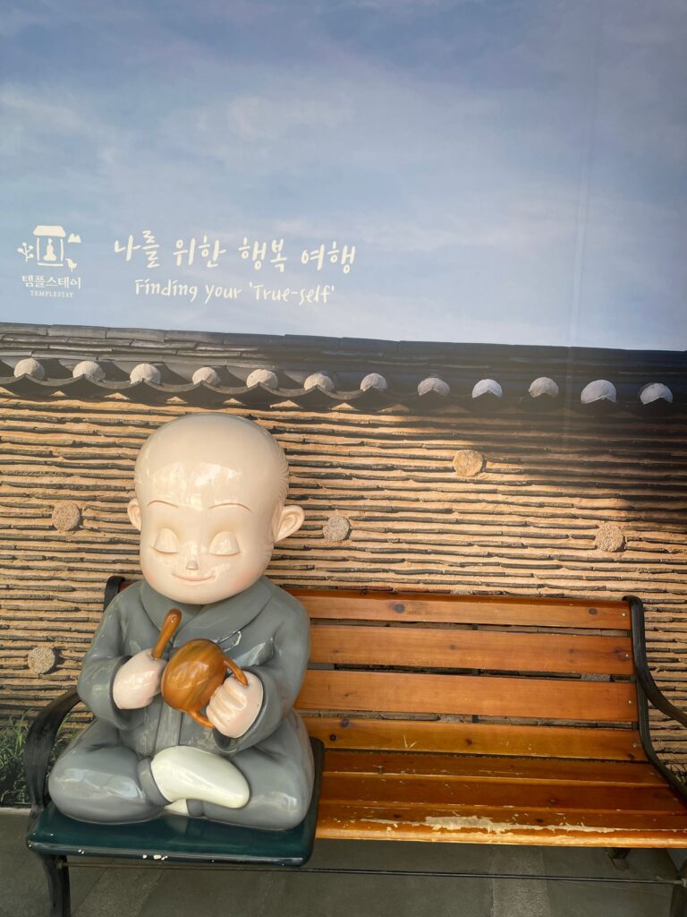 A statue of a monk on a bench near Jogyesa Temple in Seoul. There is an empty space alongside him where you can sit and take pictures. Above the monk is some Korean writing alongside the English translation "finding your true self"