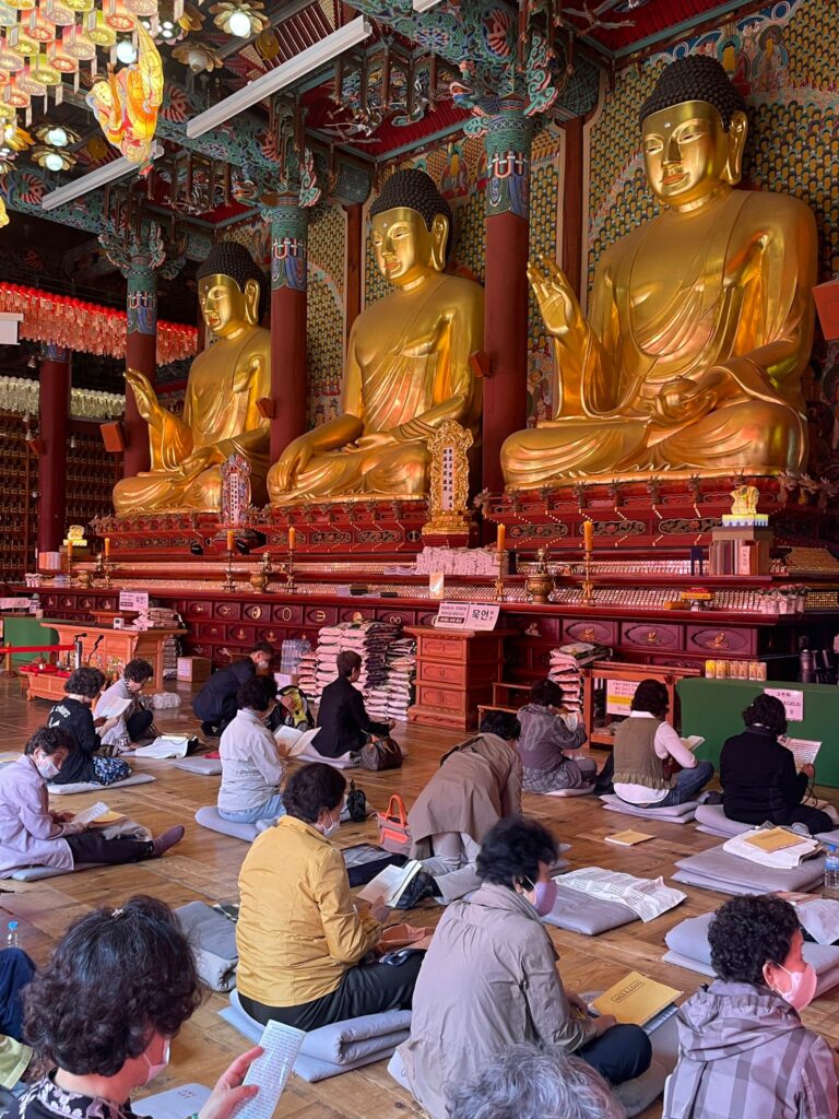 Three giant gold Buddha statues at Jogyesa Temple in Seoul. In front of them are several Koreans reading and praying whilst sat on prayer mats
