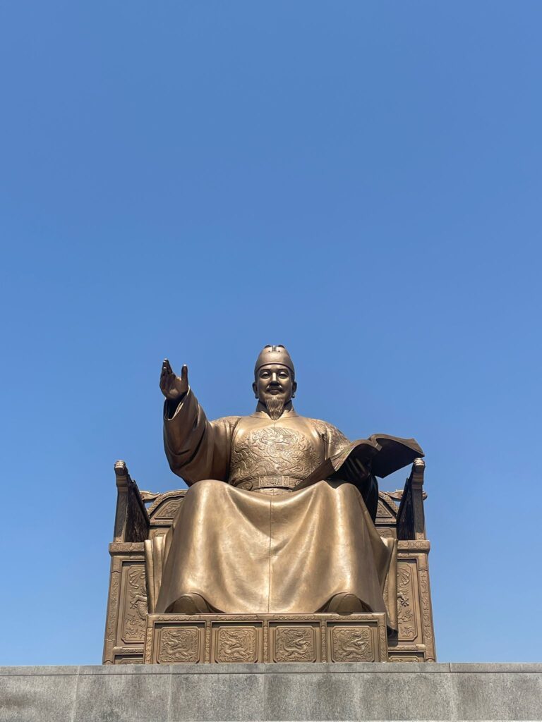 A golden statue of King Sejong the Great in Seoul, South Korea. He is sat with one hand raised and the other holding a book