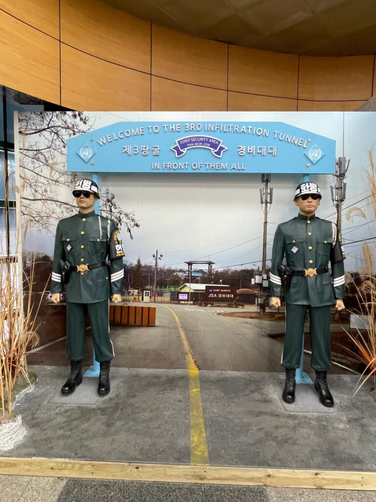 Two men in South Korean military uniform wearing glasses whilst standing watch in front of a mock-up JSA and a sign that says "Welcome to the 3rd infiltration tunnel - in front of them all"