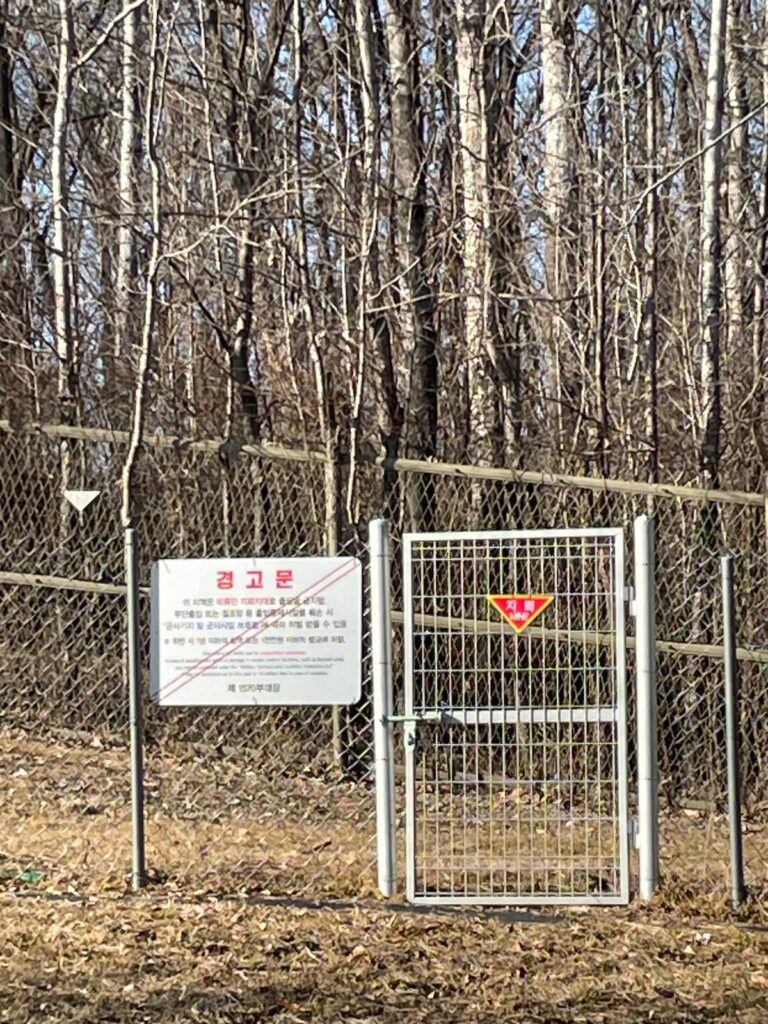 A chain fence with a red warning sign on the front which says "MINE" to warn of landmines on the other side of the fence