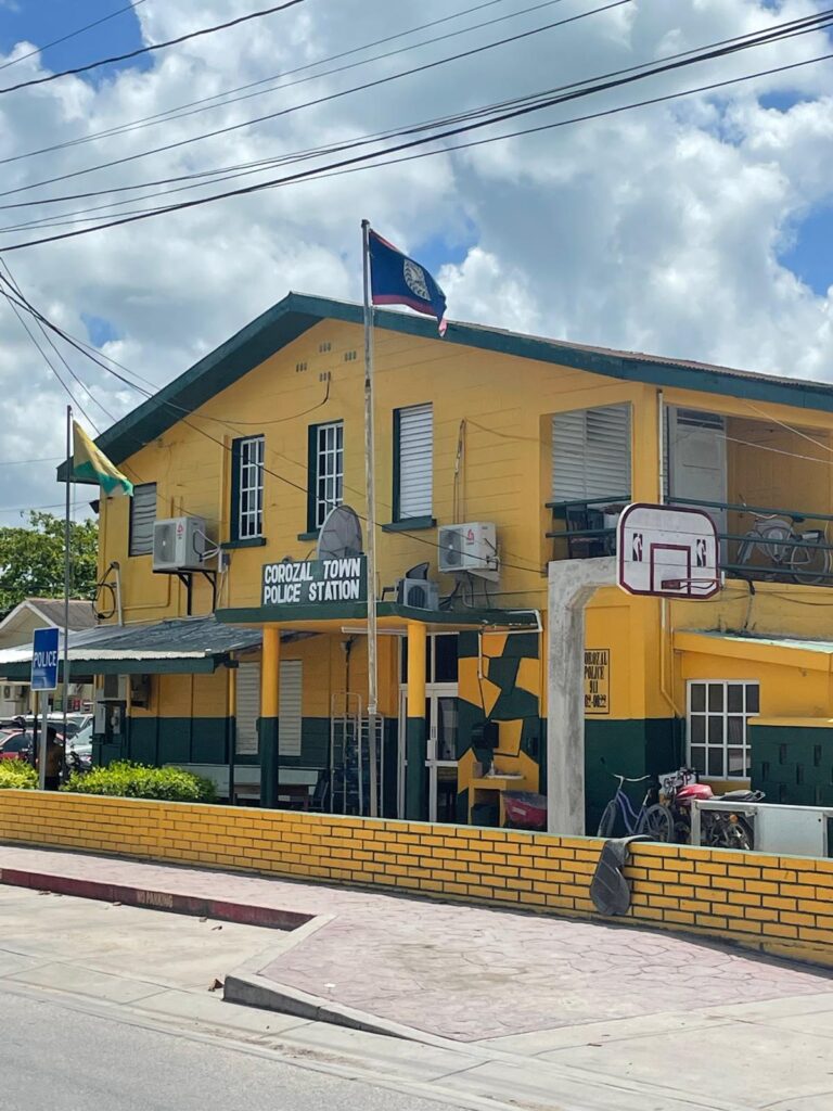 The Corozal Town Police Station building in Corozal, Belize. This dull yellow building has the Belizian flag outside along with a basketball hoop and some bicycles