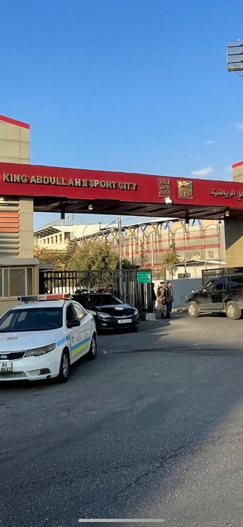 The entrance to the King Abdullah II Sport City stadium in Amman, Jordan. A police car and two other vehicles can be seen, with a police officer seen guarding the entrance
